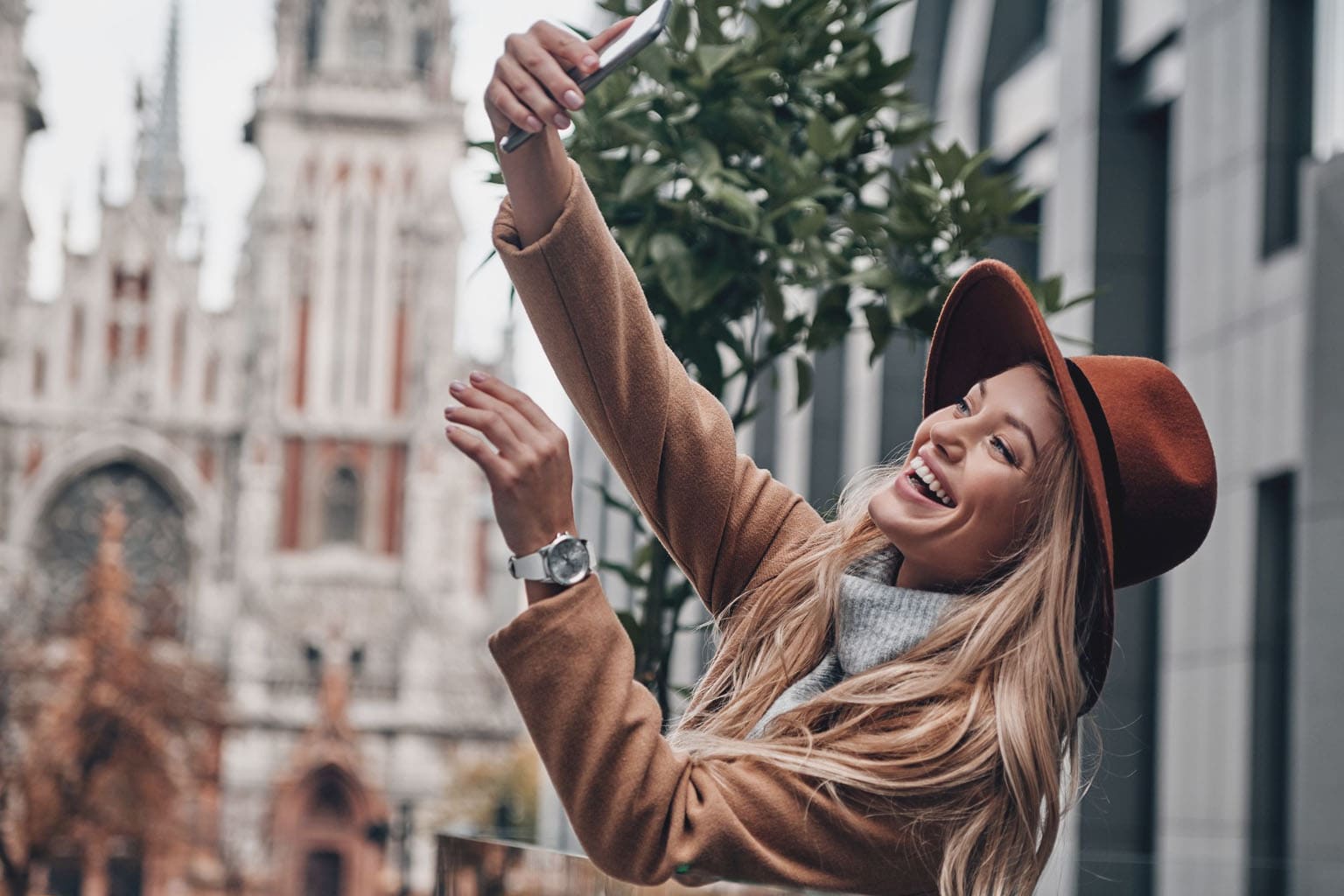 Femme souriante prenant un selfie en ville avec un chapeau, illustration d’une maman hybride moderne qui assume ses différentes vies entre maternité, travail et passions.