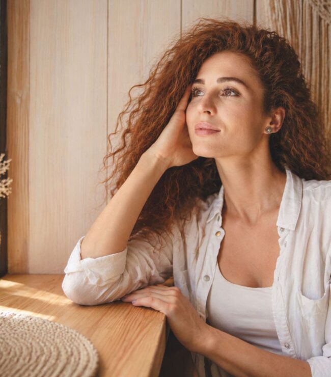 Femme pensive regardant par la fenêtre, incarnation de la charge mentale d’une maman active au quotidien