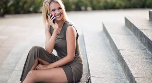 Femme blonde souriante assise sur des marches en robe kaki, téléphone à la main, incarnant une maman quadra moderne, féminine et indépendante dans un article sur la slow maman et la femme fatale.