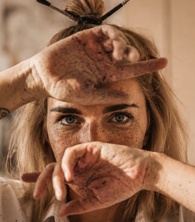 Femme de 40 ans cachant partiellement son visage avec ses mains tachées de peinture, symbole de fatigue mentale et d’auto-effacement féminin