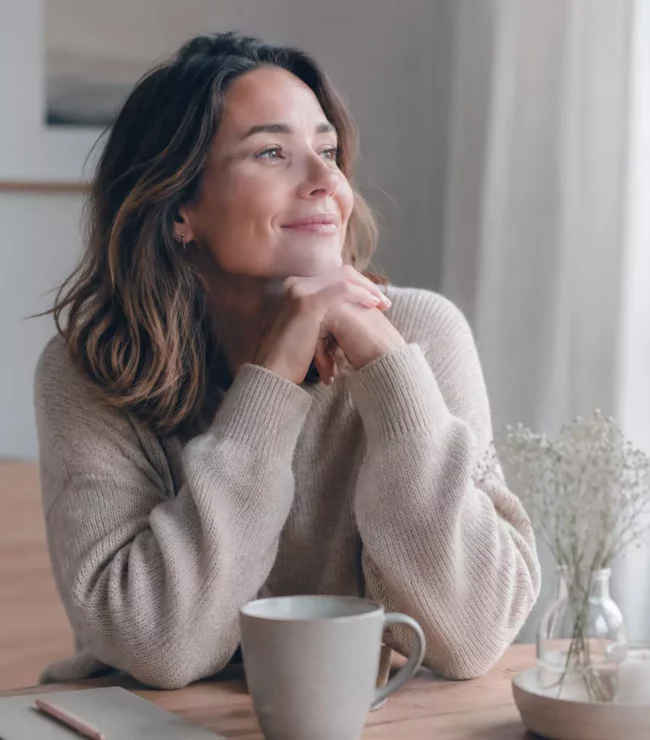 Femme de 35 ans assise près d'une fenêtre avec une tasse chaude et un carnet, prenant un moment de gratitude et de réflexion dans une ambiance slow living apaisante.