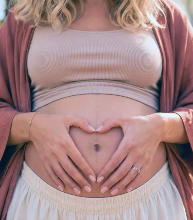 Femme enceinte aux cheveux blonds bouclés formant un cœur avec ses mains sur son ventre, symbole de la maternité choisie et du désir de devenir maman à 30 ans.