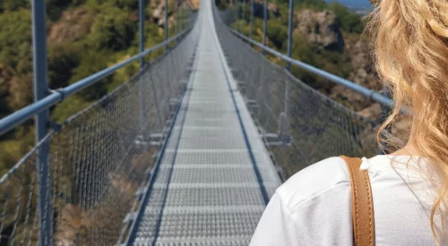 Vue de dos d'une femme aux cheveux bouclés traversant la passerelle de Mazamet en direction du village médiéval de Hautpoul, avec un panorama sur la vallée de l'Arnette.