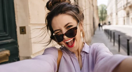Femme de 35-40 ans riant en prenant un selfie dans la rue, lunettes de soleil baissées