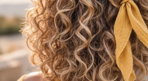 Gros plan sur des cheveux bouclés blond miel aux boucles naturelles, légèrement irrégulières, avec un foulard jaune noué dans la chevelure