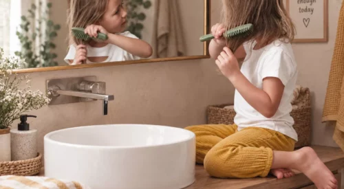 Jeune fille assise dans une salle de bain bohème, pantalon jaune moutarde et brosse verte à la main, se coiffant devant un miroir dans une routine familiale du matin