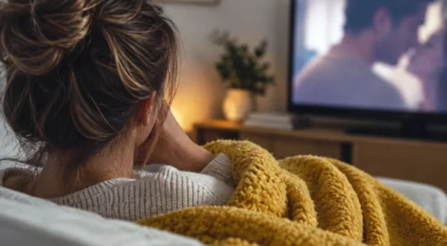 Femme de dos sous un plaid jaune regardant à la télévision une scène de couple floutée dans un salon chaleureux