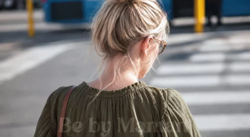 Femme blonde de dos avec chignon flou, lunettes écaille et robe vert olive dans une rue ensoleillée.